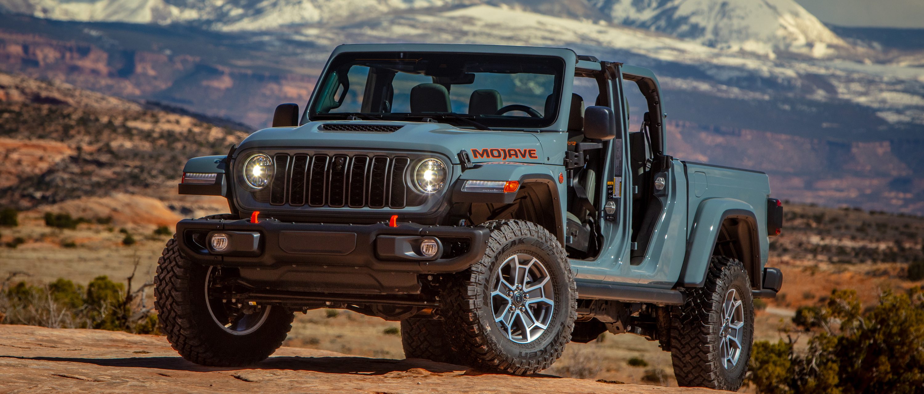 2026 Jeep Gladiator Mojave X pickup with open cabin parked on red rock with distant snow‑capped mountains, promotional photo for Leavens Chrysler London Ontario.