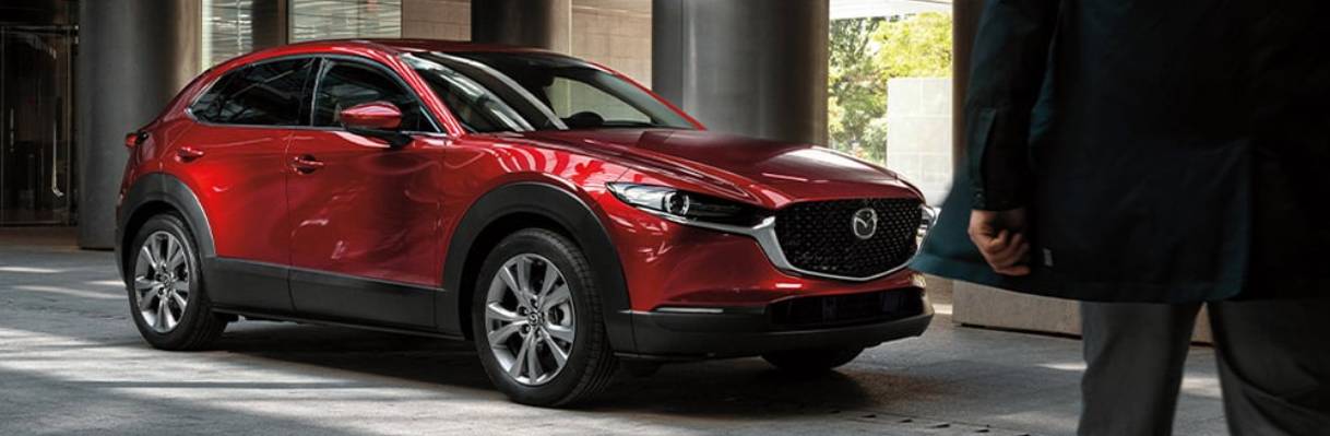 a man walks from the camera's POV towards a red 2025 Mazda CX-30 that is parked in front of some pillars