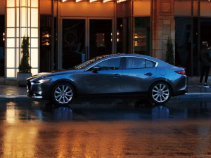 A deep blue 2025 Mazda3 is parked outside of a high-class hotel in the city. The windows of the hotel lobby glow an inviting yellow. The road under the Mazda3 is wet and reflects the windows and the evening sky.