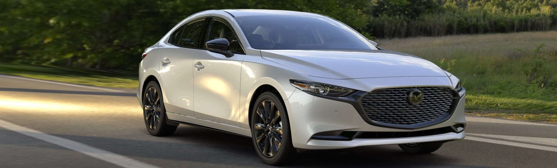 A silver 2025 Mazda3 drives at an angle towards the camera. Greenery can be seen by the side of the road.