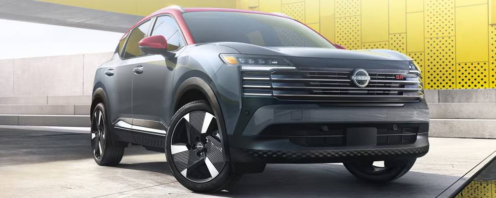 A grey 2026 Nissan Kicks with a red roof is parked under a bridge in front of a concrete wall in a city. The wall alongside it is painted a bright yellow