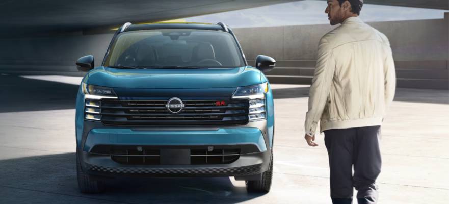 A blue 2026 Nissan Kicks is parked in a covered parking lot. A man in a white jacket walks towards it