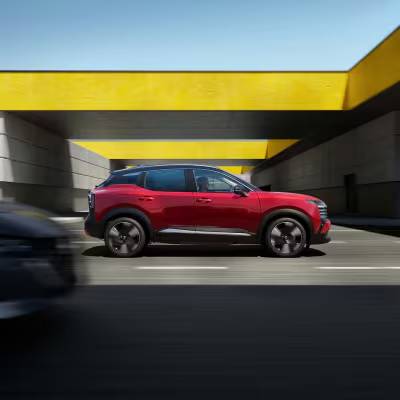 A red 2026 Nissan Kicks drives past a concrete structure with a yellow roof