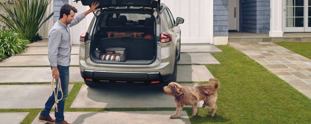 A man and a dog stand in front of a 2026 Nissan Rogue's open trunk. Why the Nissan Rogue is the Perfect Road Trip Vehicle at Medicine Hat Nissan in Medicine Hat, AB