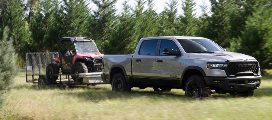A silver 2026 Ram 1500 pulls a red vehicle on a trailer across a field. New 2026 Ram 1500 for sale at Orangeville Chrysler Dodge Jeep Ram in Orangeville, ON