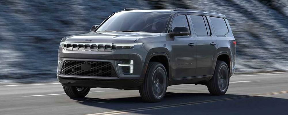 A dark grey 2026 Jeep Grand Wagoneer drives past the camera in front of a cliff face