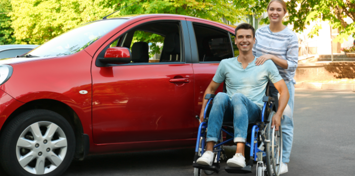 person_standing_behind_person_in_wheelchair_next_to_red_car_parked_in_parking_lot