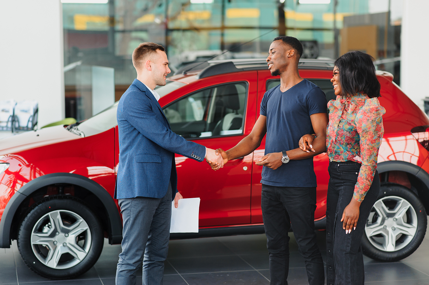 Two people shaking hands with a third person also present. All three are standing near a vehicle