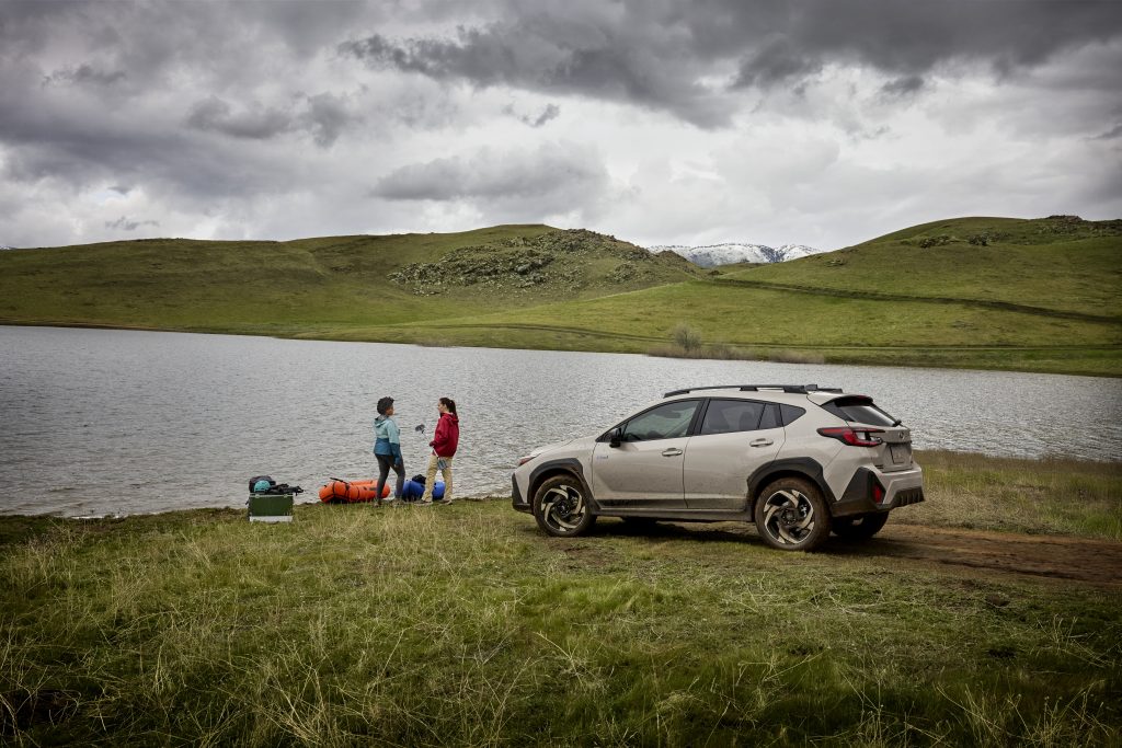 2026 Subaru Crosstrek parked on grass beside a lake while two people stand near camping gear on the shoreline under cloudy skies.