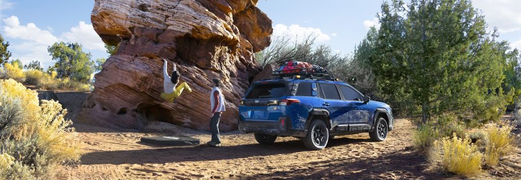 Blue 2026 Subaru Outback Wilderness parked on sandy terrain near rock formations with outdoor gear secured on the roof.