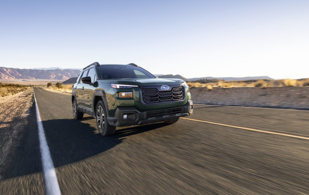 Green 2026 Subaru Outback SUV driving on a desert highway with mountains in the background.