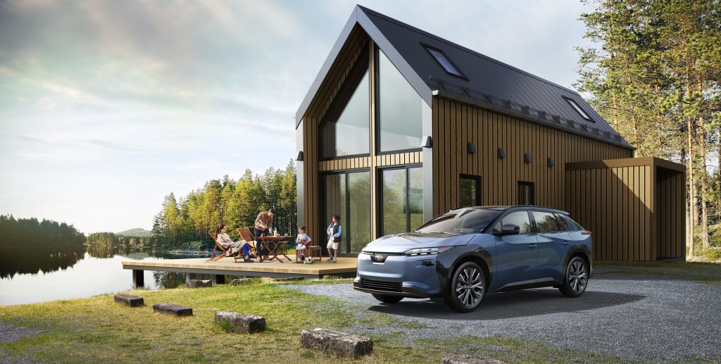 Blue 2026 Subaru Solterra parked in front of a wood‑clad cottage by a lake, with a family relaxing on the deck nearby.