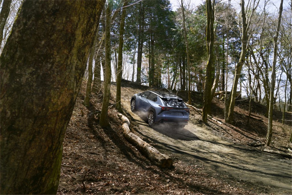 Blue 2026 Subaru Solterra driving up a dirt incline through a dense forest, kicking up dust on the trail.
