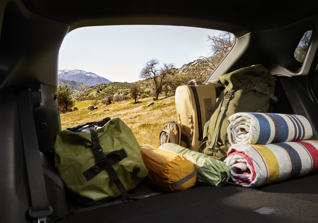 Rear cargo area of a 2026 Subaru Solterra filled with duffel bags, backpacks, tents and rolled blankets with a scenic mountain landscape outside.
