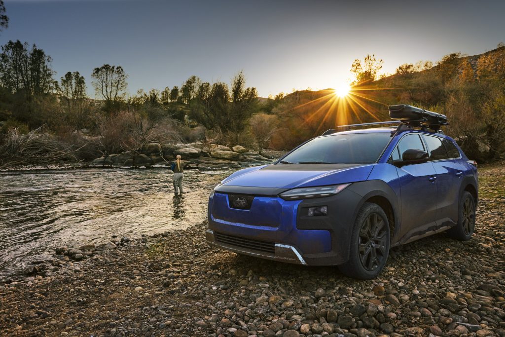 Blue 2026 Subaru Trailseeker parked on a rocky riverbank at sunset with a person fly‑fishing in the water nearby.