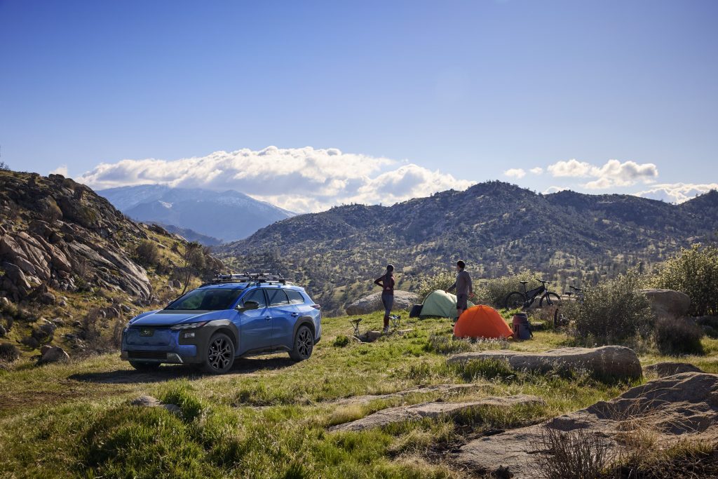 Blue 2026 Subaru Trailseeker parked on a grassy hilltop near colourful tents, people and mountain bikes with distant mountains under a clear sky.