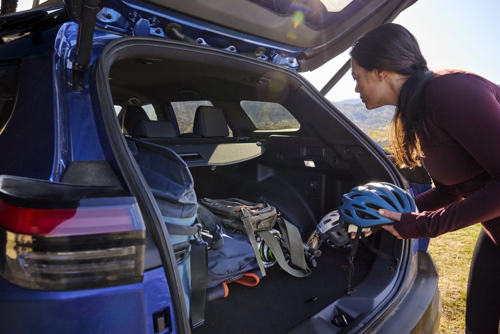Woman loading helmets and outdoor gear into the open rear cargo area of a blue 2026 Subaru Trailseeker with the liftgate raised.