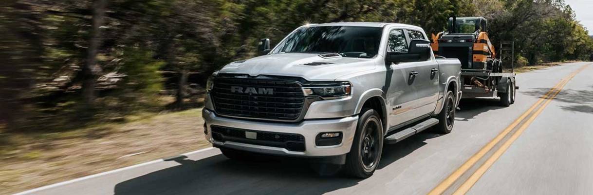 A silver 2026 Ram 1500 pulls a trailer bearing a vehicle on a road bordered by trees