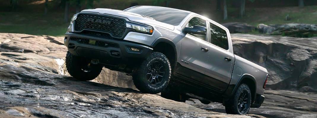 A silver 2026 Ram 1500 sits on a dark rock slope, tilted up
