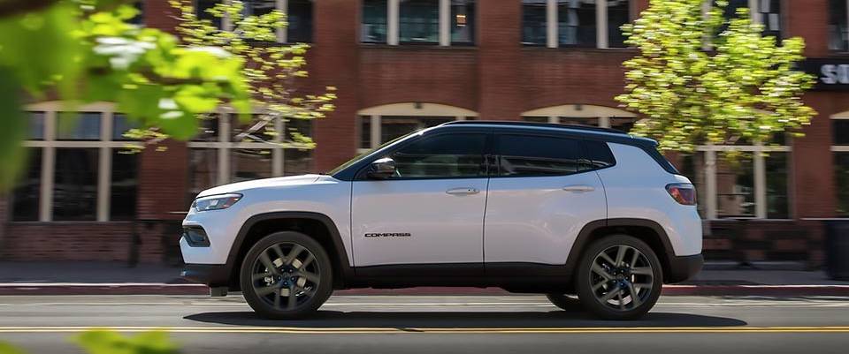 A white 2026 Jeep Compass drives past a building and trees
