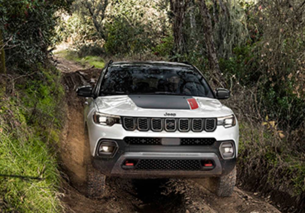 A white 2026 Jeep Compass with a black hood spoiler drives through a forest