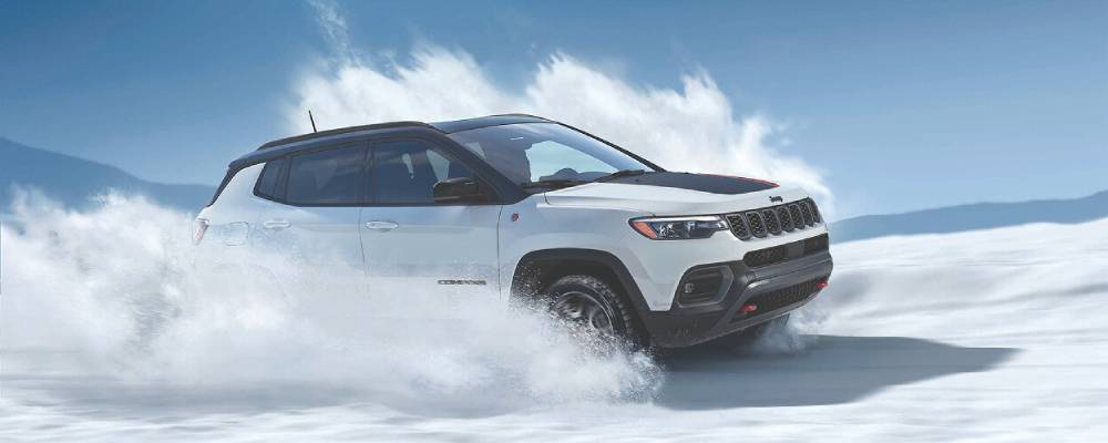 A side-on view of a white 2026 Jeep Compass with a black and red roof decal drives past the camera kicking up snow