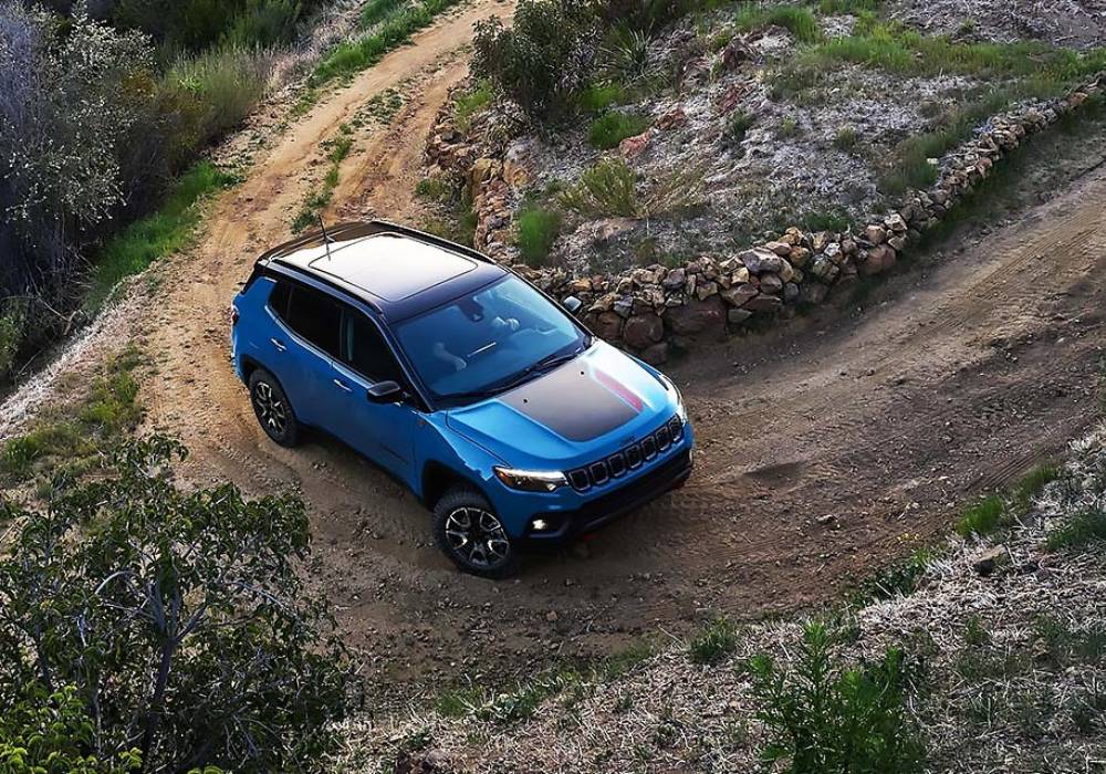 A blue 2026 Jeep Compass with a black hood decal takes a curve on a dirt road
