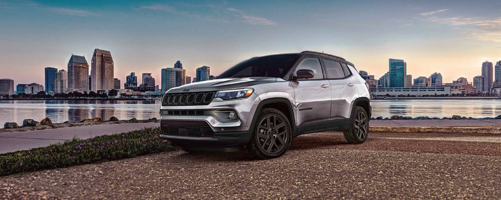A silver 2026 Jeep Compass is parked in front of a harbour with a city skyline on the other side