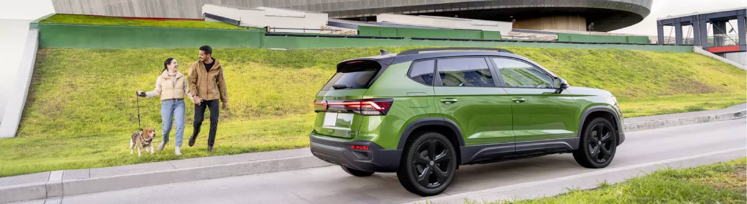 A green 2025 Volkswagen Taos passes a dogwalking couple on a road with a green hill beside it. A circular building can be seen in the background.