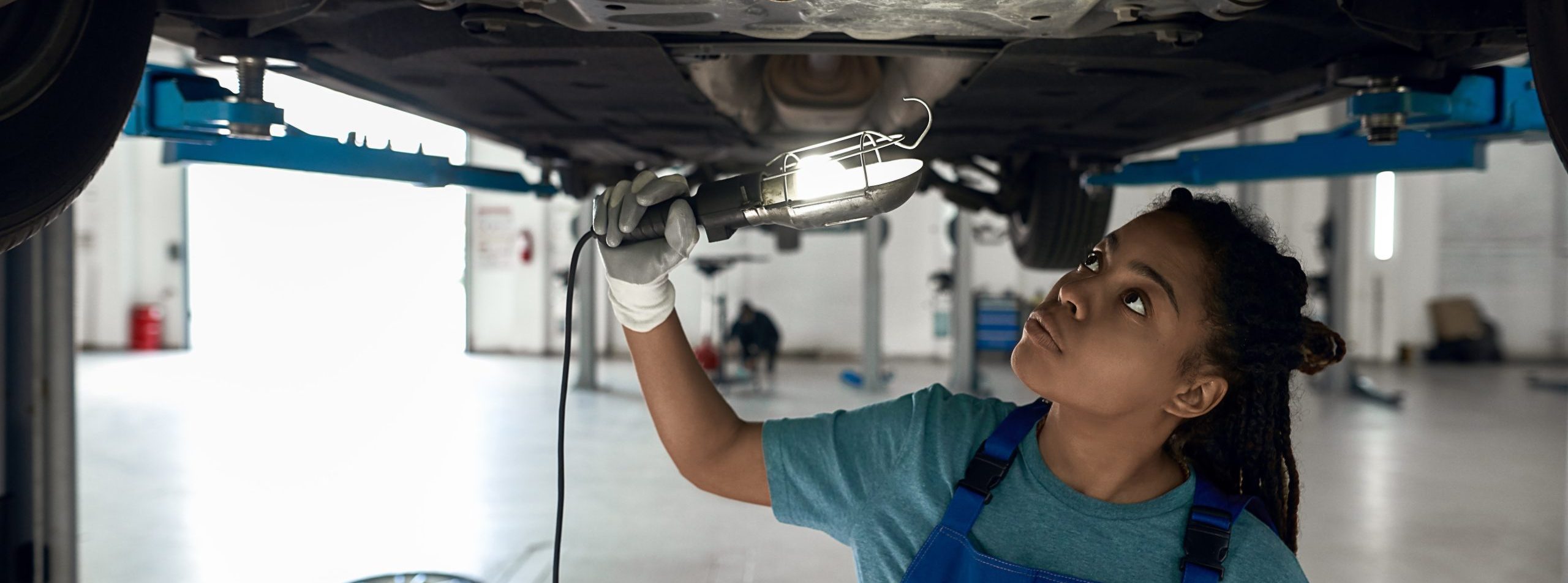 Young afro american girl checking bottom of car installed on lift at car service station with flashlight . Auto repairing concept