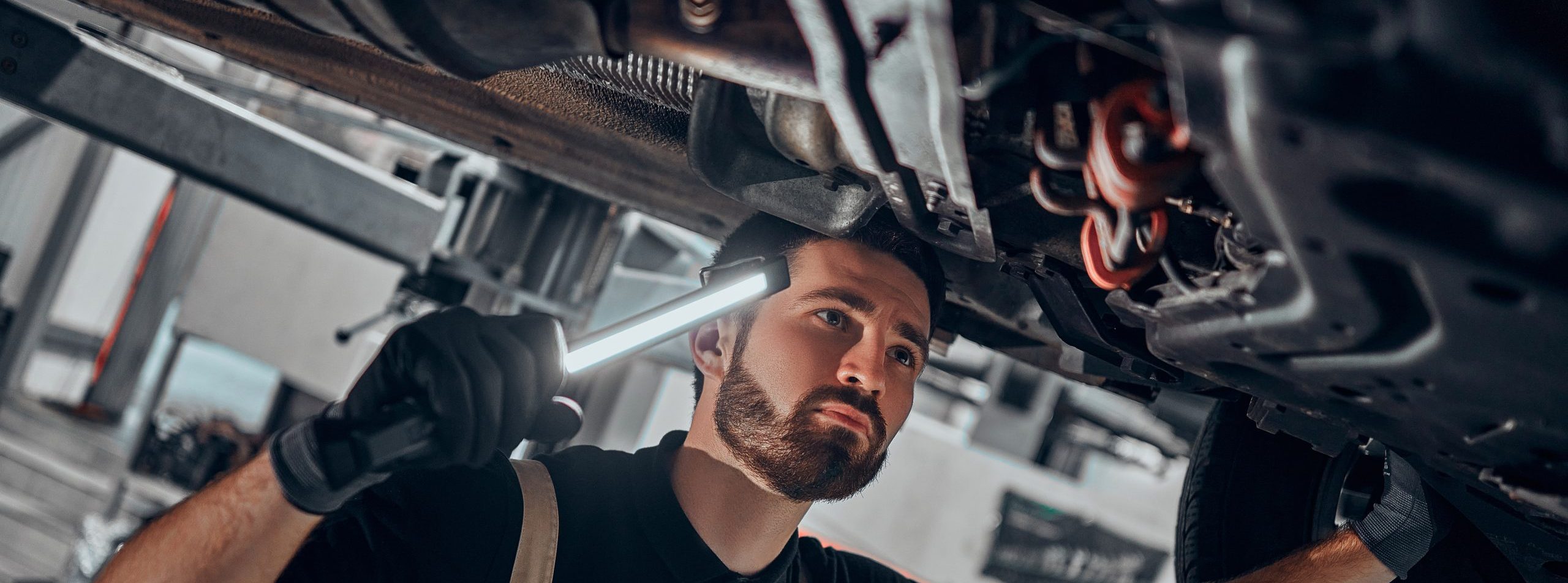 Portrait shot of a handsome mechanic working on a vehicle in a car service. Professional repairman is wearing gloves and using a lamp underneath the car. Modern clean workshop.