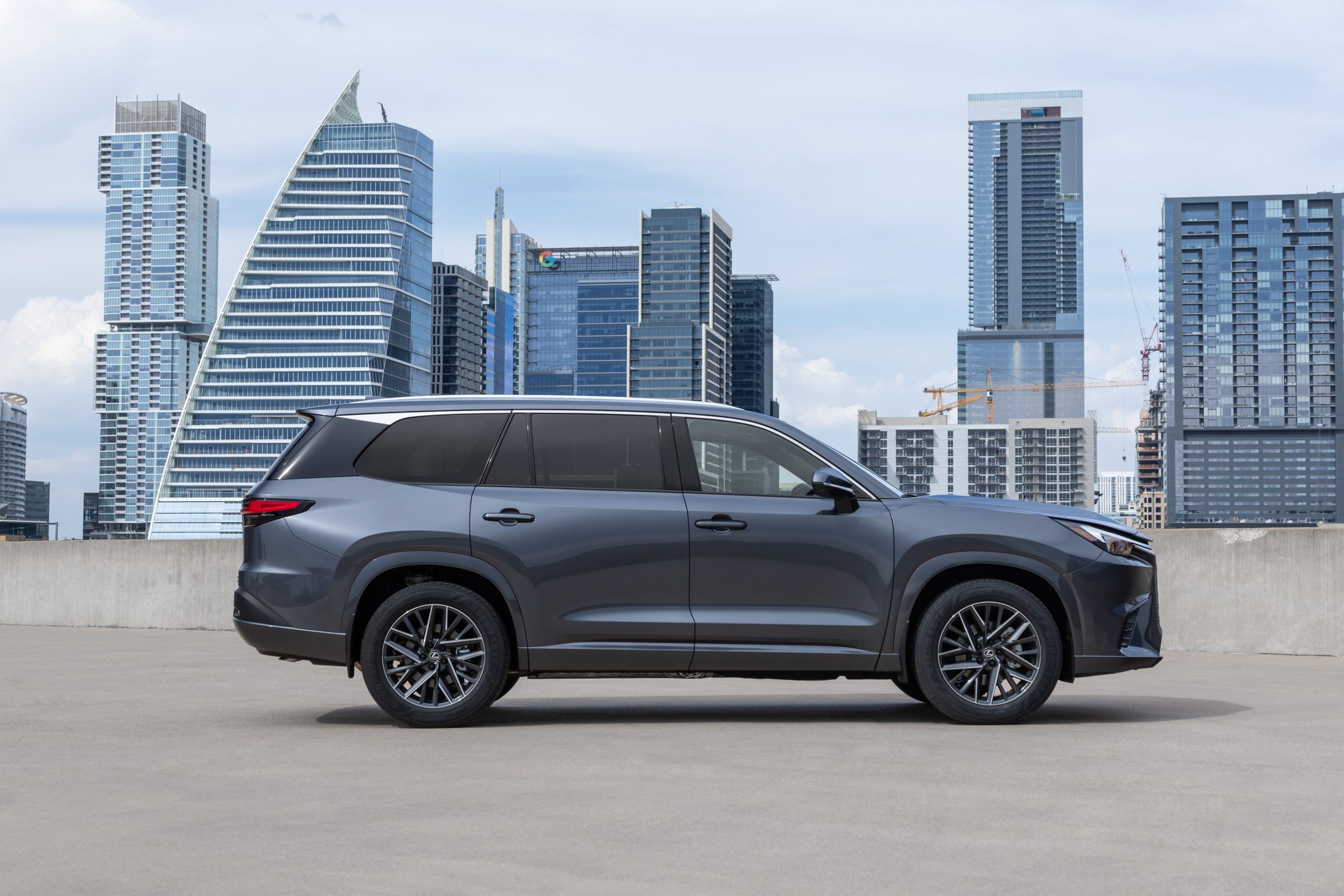 Side profile of a 2026 Lexus TX 350 Ultra Luxury in Nightfall Mica, parked on a rooftop with modern city skyscrapers in the background.