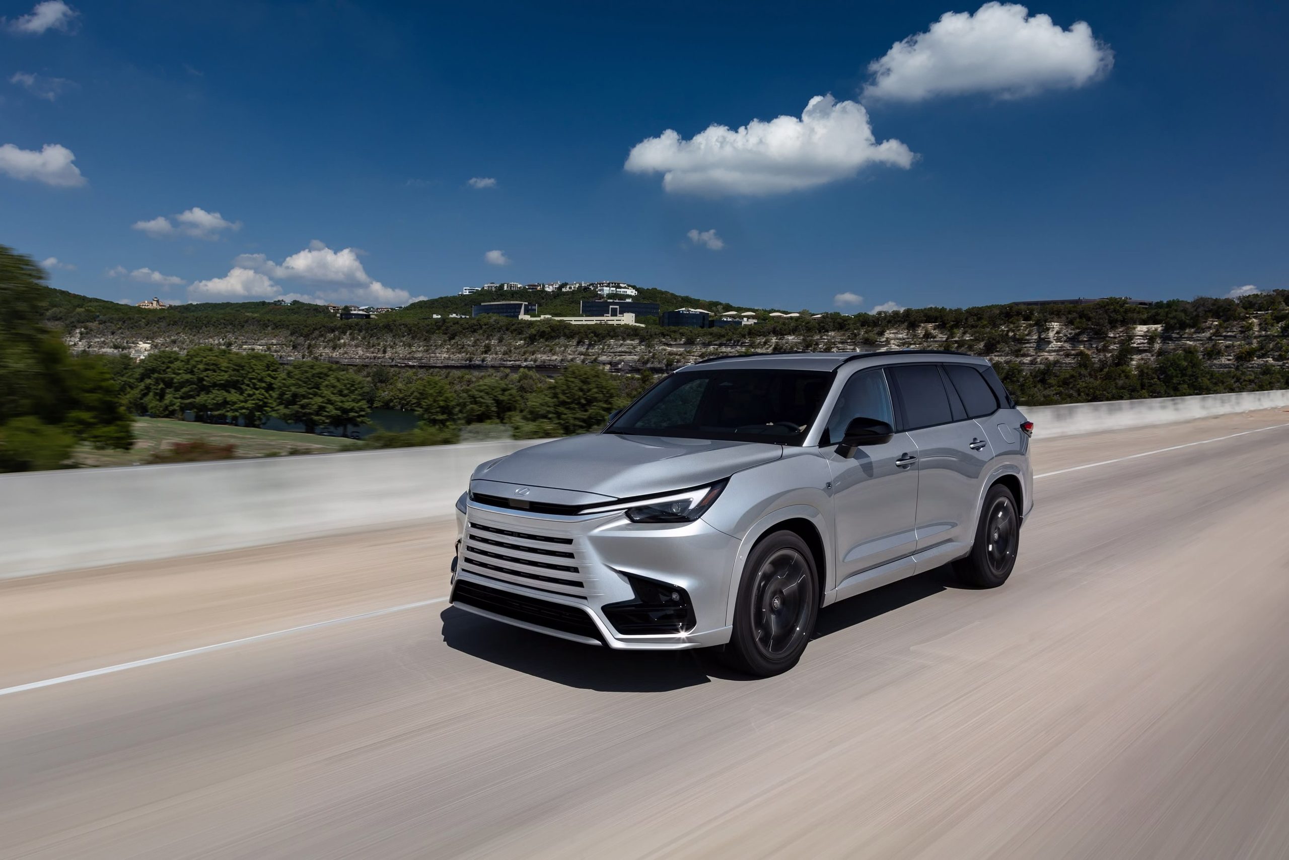 2026 Lexus TX 500h F SPORT Performance in Celestial Silver Metallic driving on a scenic highway under a bright blue sky.