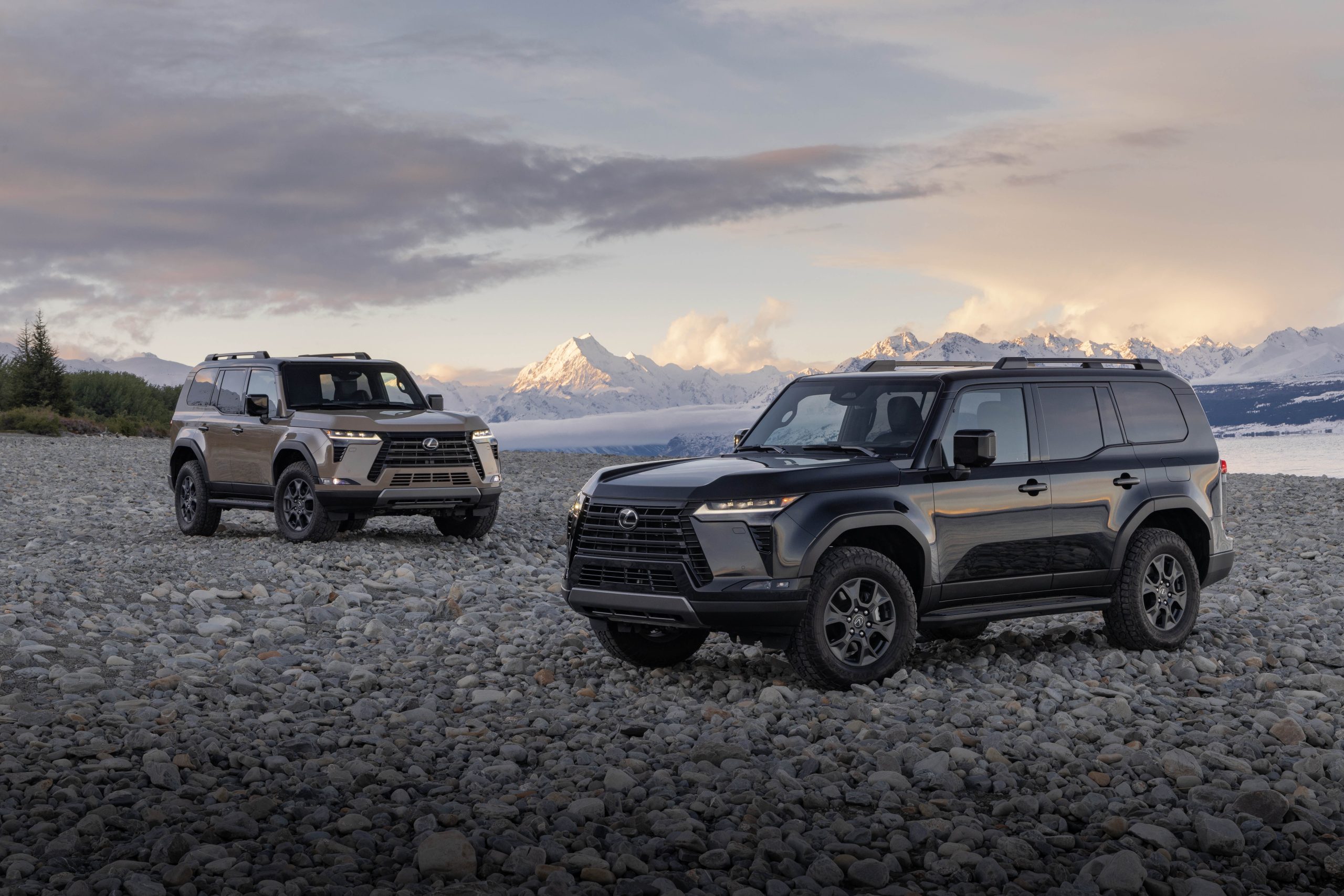 Two 2026 Lexus GX 550 Overtrail models parked on a rocky shoreline with snow-capped mountains in the background at sunrise, showcasing their rugged off-road design.