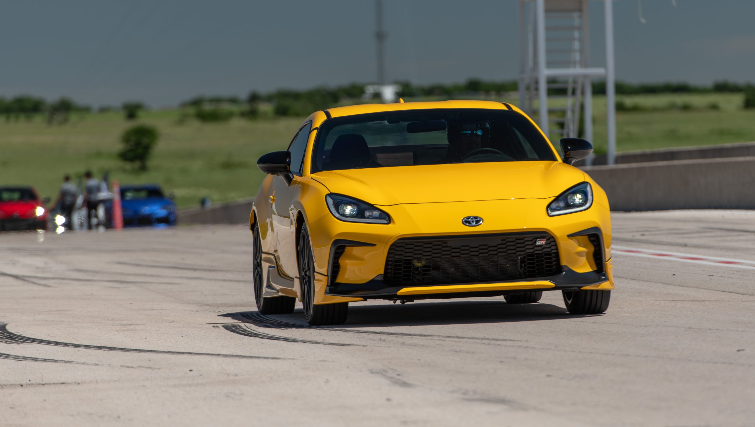 2026 Toyota GR86 Yuzu Edition in vibrant yellow driving on a racetrack, showcasing its sporty stance, aggressive front grille, and performance-focused design.