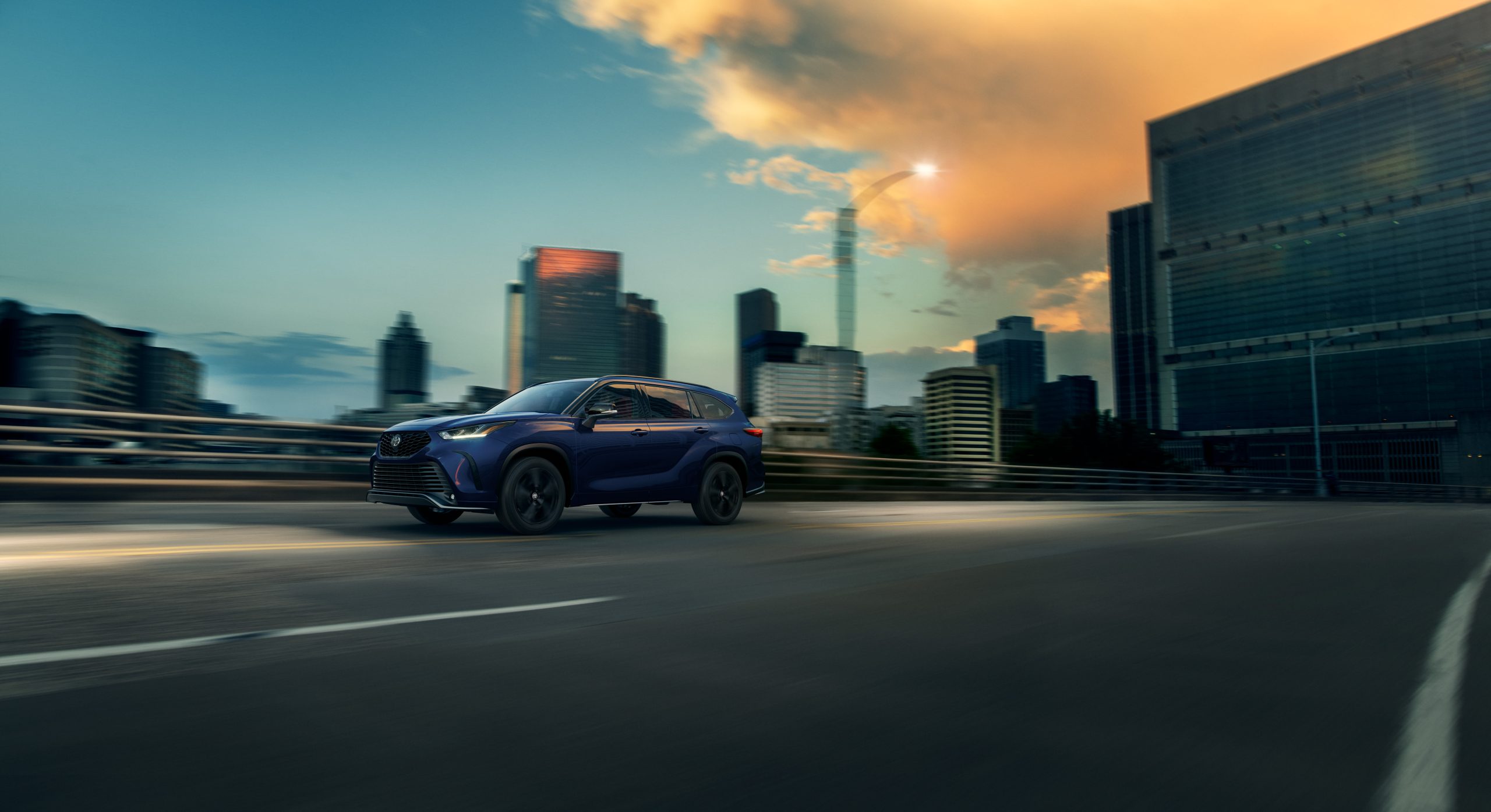 2026 Toyota Highlander XSE in Blueprint driving through the city at sunset, showcasing its sporty design and bold black alloy wheels.