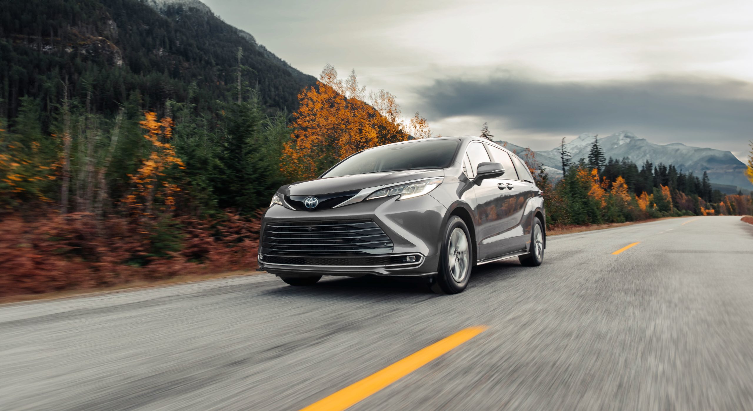 Front view of a grey 2026 Toyota Sienna Hybrid LE driving on a mountain highway with fall trees.