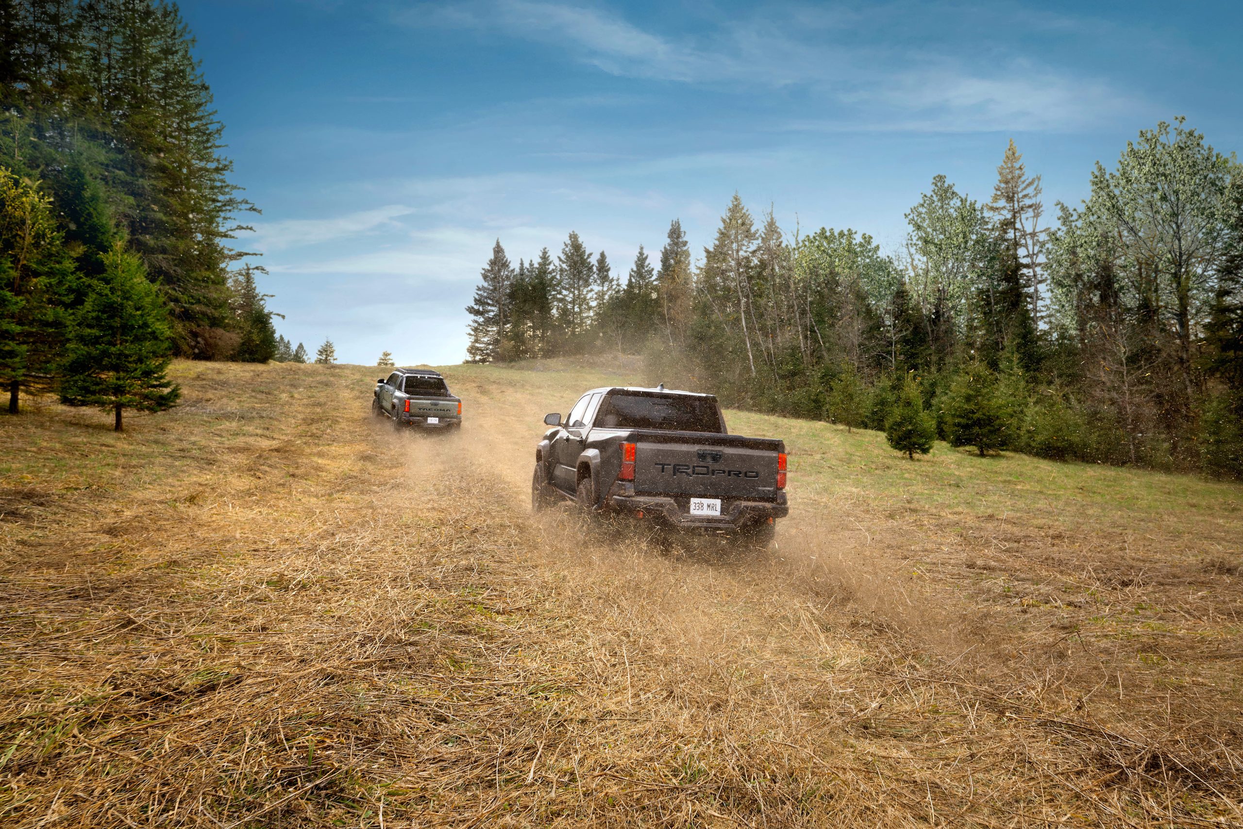 Two Toyota Tacoma TRD Pro pickup trucks driving across a grassy field near a forest
