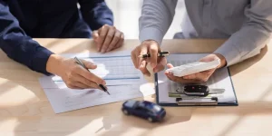 A man points at paperwork being signed by a driver for a used vehicle with a toy car on the table.