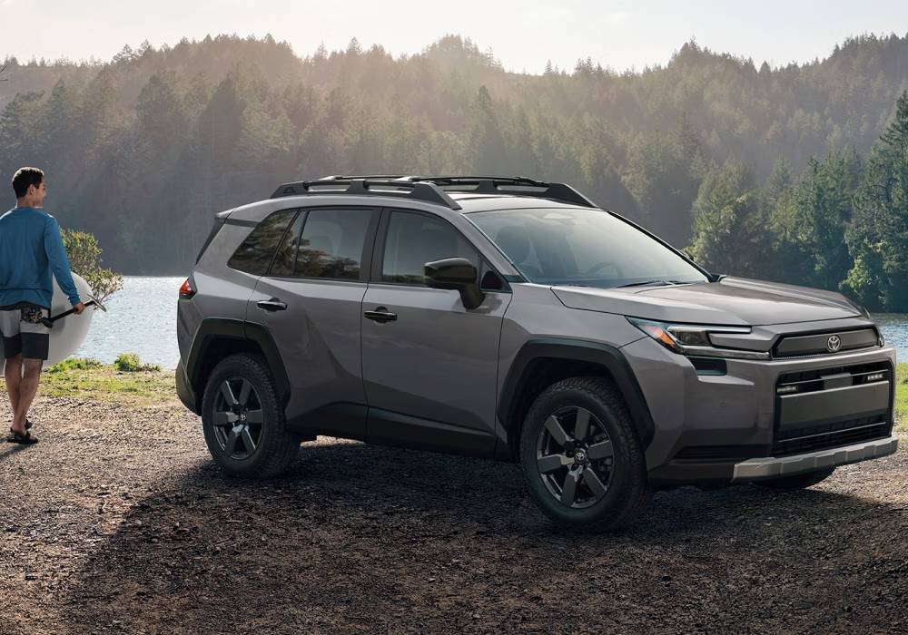 A silver 2026 Toyota RAV4 is parked in front of a lake with trees in the background